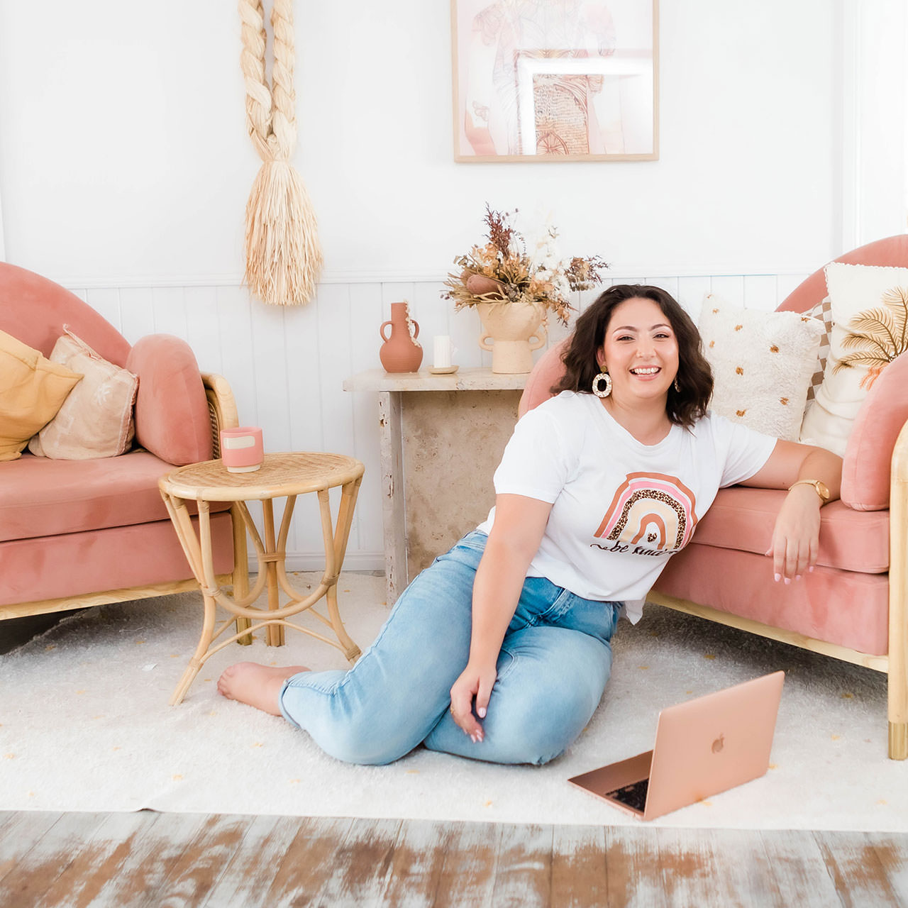 Woman sitting on the floor with a laptop, surrounded by cozy pink furniture and decor.